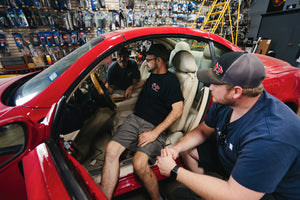 Three men discussing car repairs inside a red sports car at an auto parts shop.