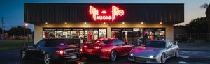 An audio store with neon signs at twilight, flanked by sports cars in the parking lot.